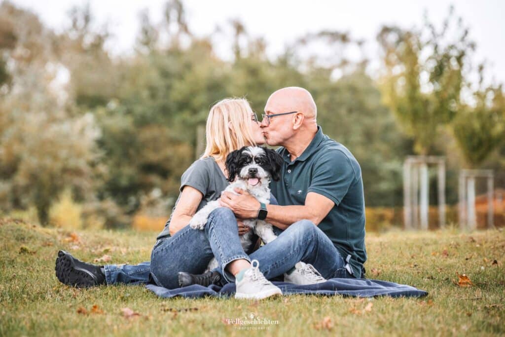 Ehepaar sitzt mit seinem Hund auf einer Wiese und küsst sich